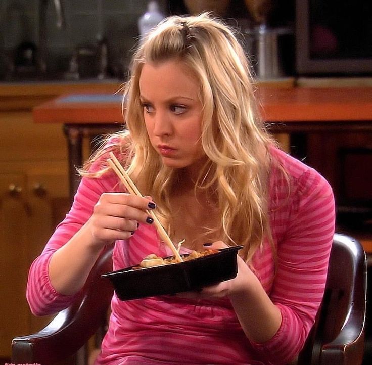 a woman sitting in a chair holding a bowl of food and chopsticks to her mouth