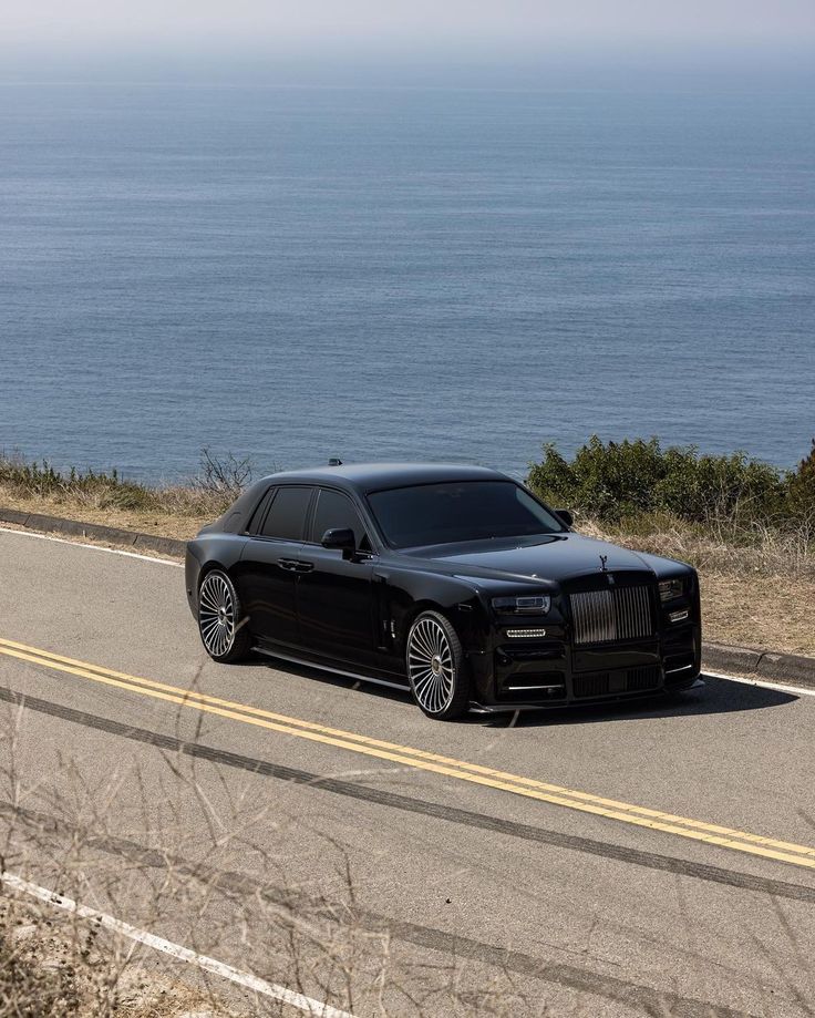 a black rolls royce parked on the side of the road next to the ocean in front of it