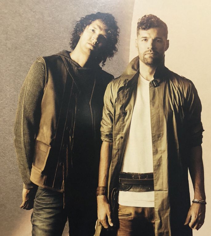 two men standing next to each other in front of a white wall and one is holding a brown bag