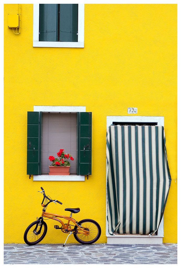 a bike parked next to a yellow building with green shutters and a striped chair