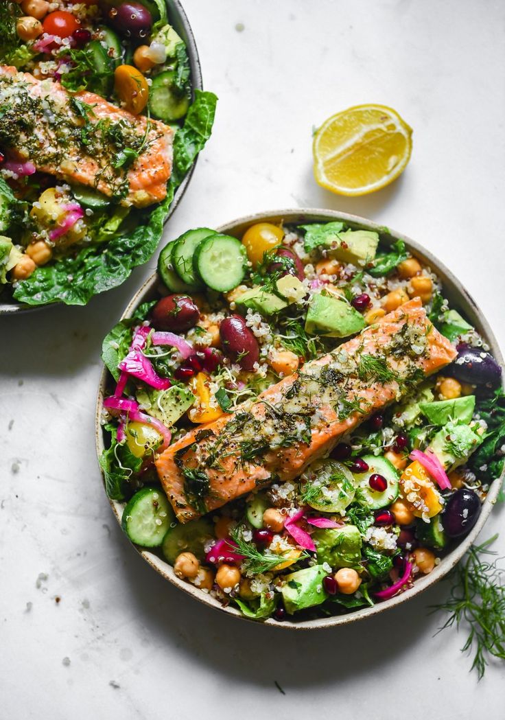 two bowls filled with salad and salmon on top of a white table next to a lemon wedge