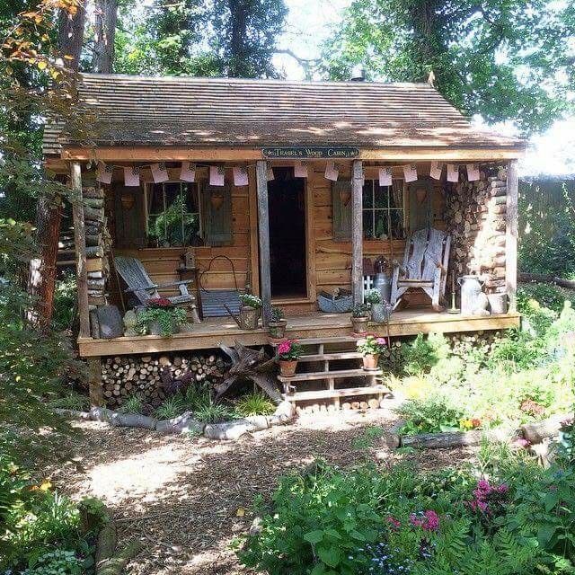 a small wooden cabin in the woods surrounded by flowers and trees with chairs on the porch