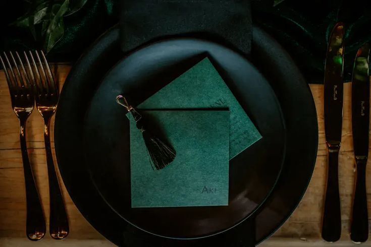 a place setting with green napkins and silverware on a black plate, surrounded by greenery