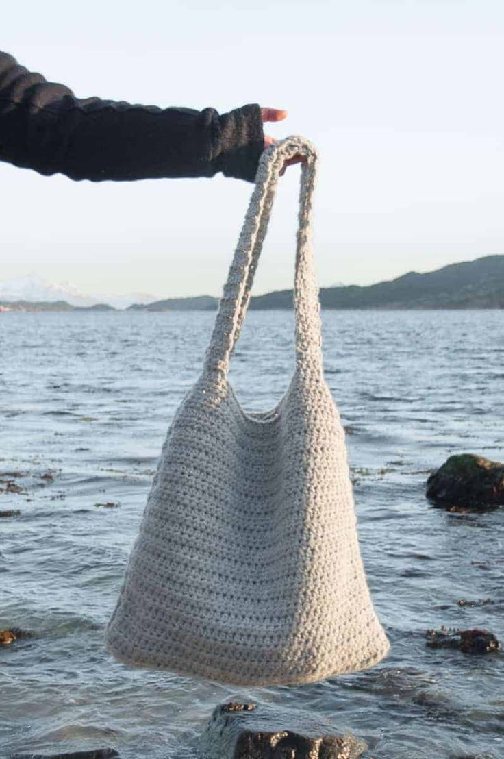 a hand holding a crocheted bag over the water's edge with rocks in the background