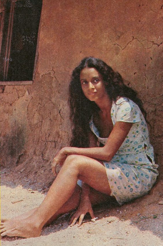 a woman sitting on the ground in front of a brown wall with a small window