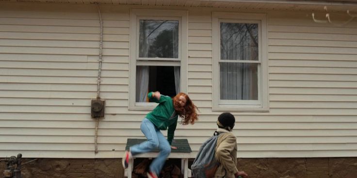 two people standing in front of a house and one person sitting on a chair outside