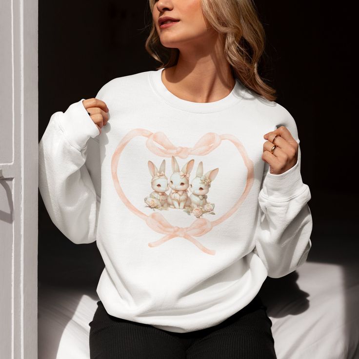 "Introducing our \"Bunny Love\" Sweatshirt - a heartwarming and whimsical piece that captures the essence of adorable bunnies and a touch of romance. This sweatshirt is more than just cozy wear; it's a wearable work of art, featuring a captivating image of three adorable bunnies nestled within a heart-shaped frame made of a soft peach-colored ribbon. Crafted with meticulous attention to detail, this sweatshirt brings the innocence and charm of bunnies to life. The pastel tones of the bunnies, al Cute White Long Sleeve Sweatshirt, Cute Heart Graphic Tops For Fall, Cute Fall Tops With Heart Graphic, Cute Long Sleeve Tops With Heart Graphic, Cute Long Sleeve Top With Heart Graphic, Pink Crew Neck Top With Bunny Design, Casual Sweatshirt With Heart Graphic For Loungewear, Cute White Crew Neck Sweater, Cute White Crew Neck Sweatshirt