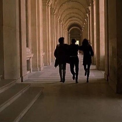 two people walking down a long hallway with columns on either side and light at the end