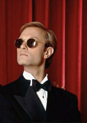 a man in a tuxedo and sunglasses standing next to a red curtain with his arm around another man's shoulder