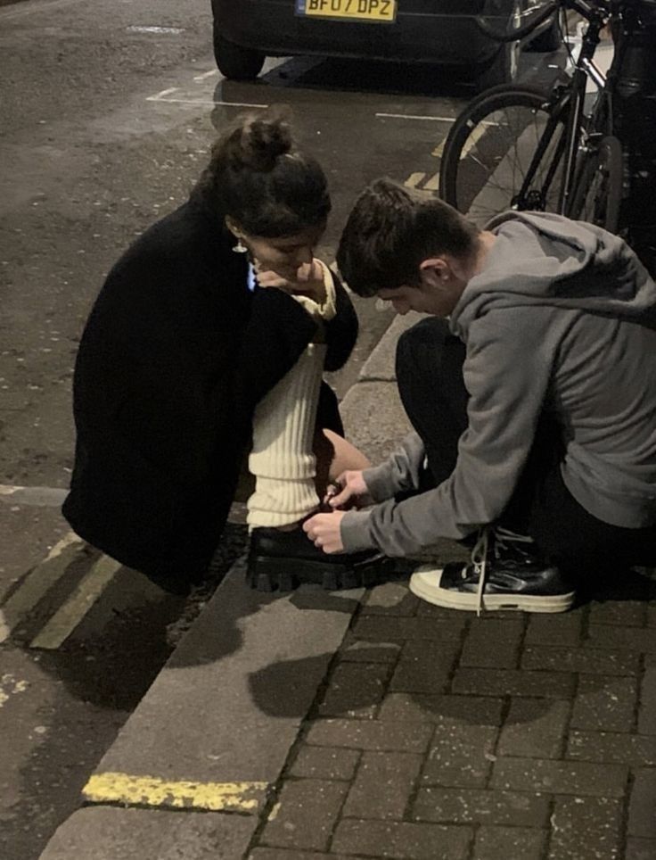 two people are sitting on the curb looking at their cell phones while one person stands over them