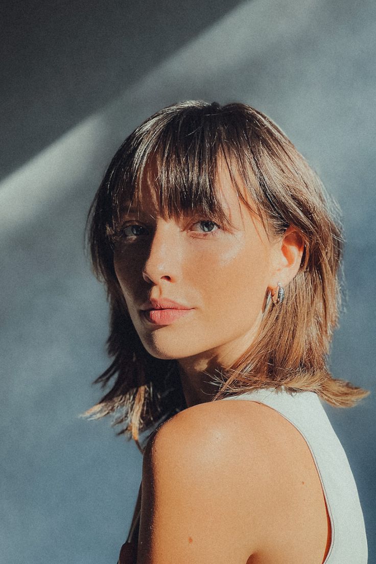 a woman with short hair wearing a tank top and looking off to the side, standing in front of a concrete wall