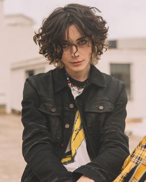 a young man with curly hair sitting in front of a white building wearing a black jacket