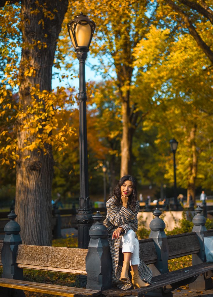 a woman sitting on a park bench next to a lamp post and tree with yellow leaves