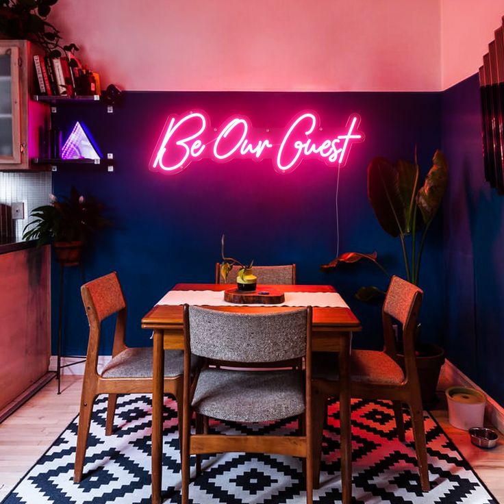 a table with chairs and a neon sign on the wall above it that says be our guest