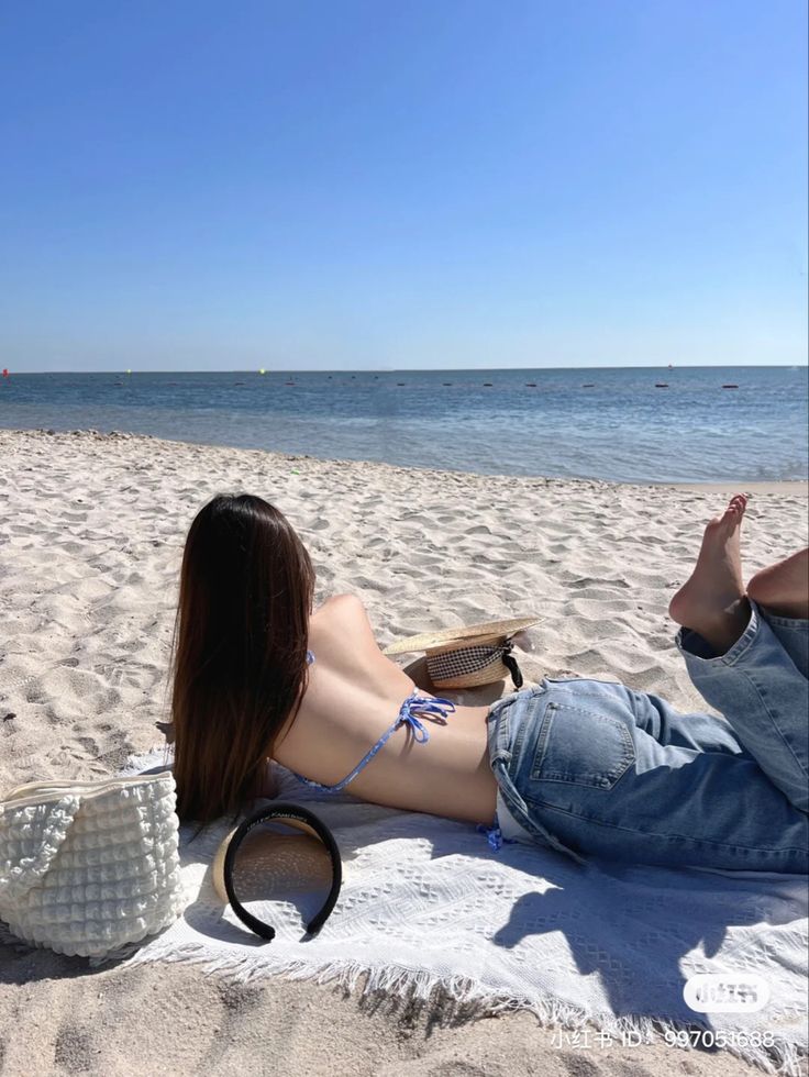 a woman laying on top of a beach next to the ocean with her legs crossed