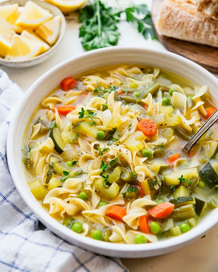 a bowl of vegetable noodle soup on a table with bread and parmesan cheese