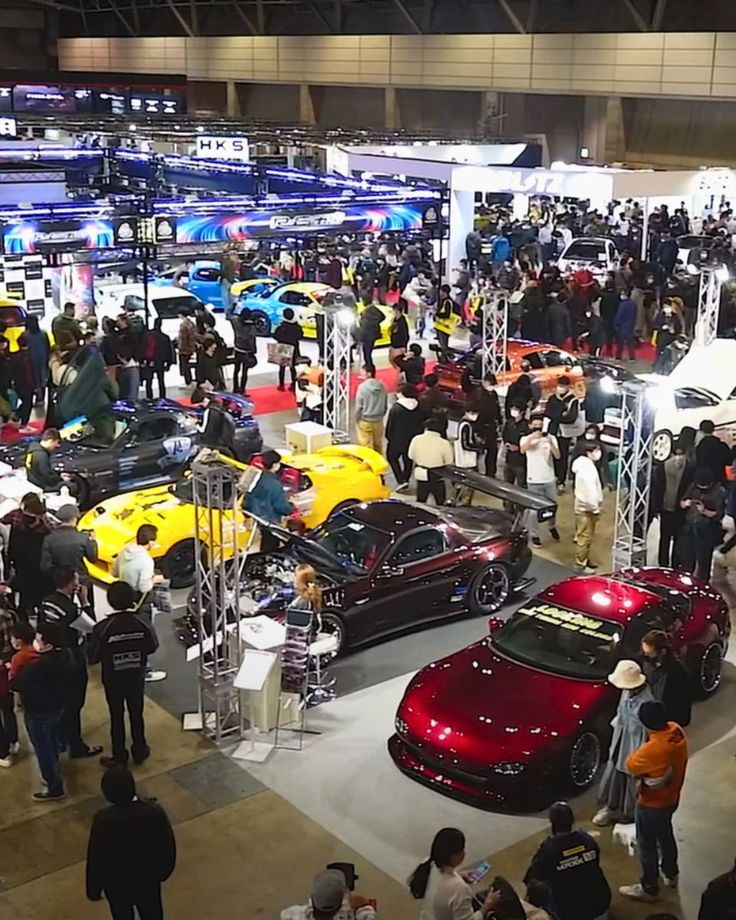 a group of people standing around cars at a car show in an indoor area with lots of vehicles on display