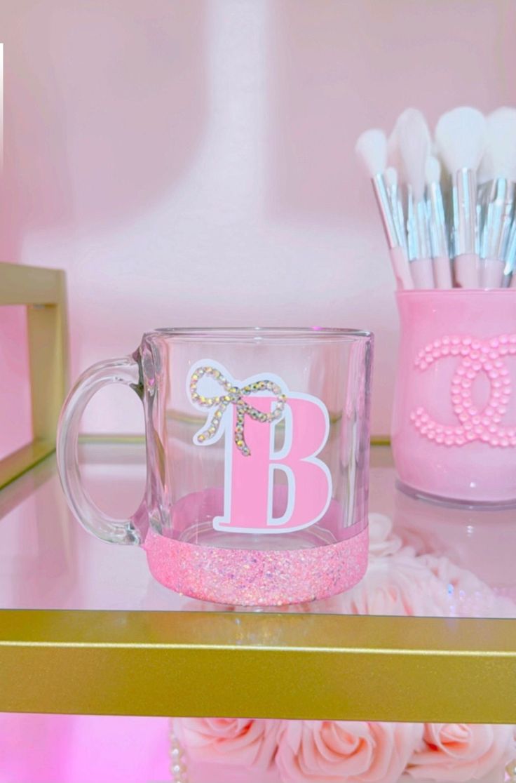 a pink and gold desk topped with a glass cup filled with makeup brushes next to a vase