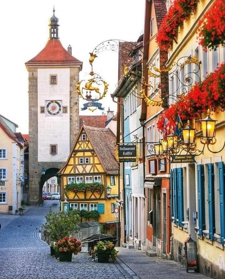 a cobblestone street lined with tall buildings and colorful flowers in front of a clock tower