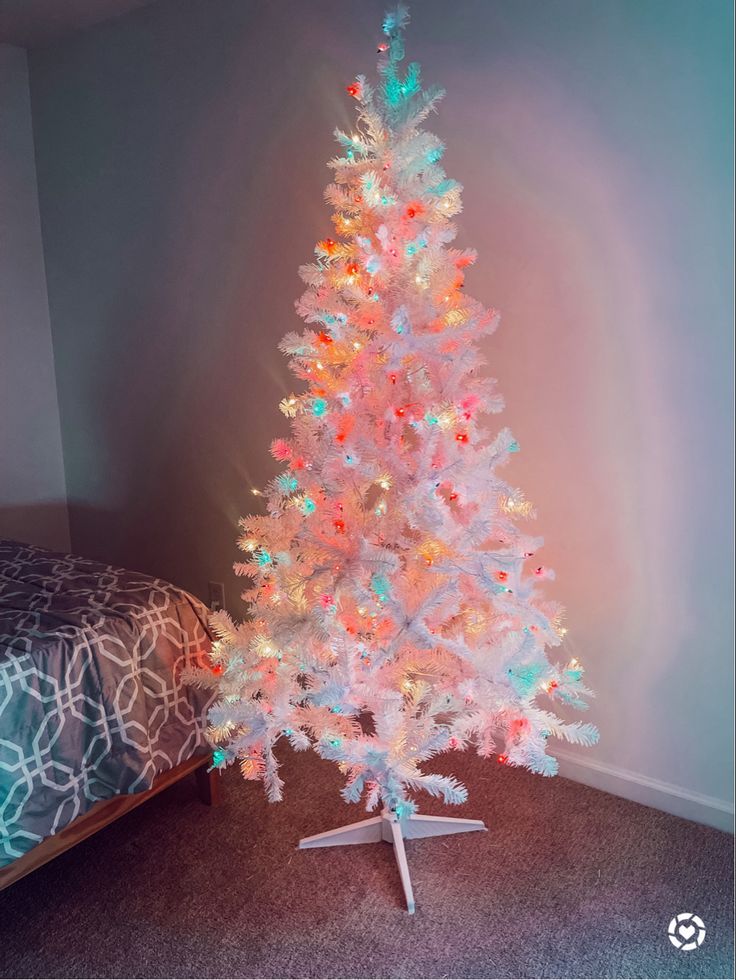 a white christmas tree with multicolored lights in a room next to a bed