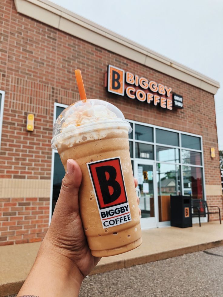 a person holding up a cup of coffee in front of a bigggy coffee shop