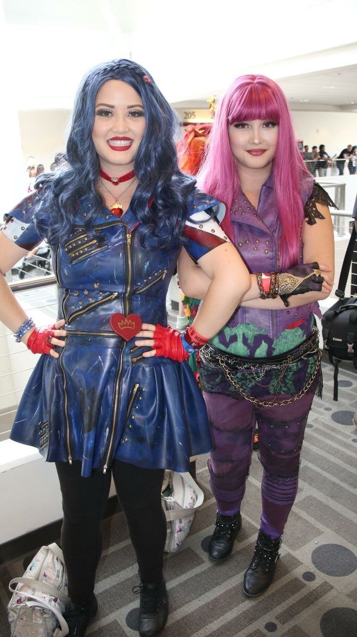 two women dressed in costumes standing next to each other on a carpeted floor, one with pink hair and the other wearing blue