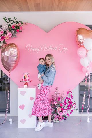 two women standing in front of a heart shaped backdrop with balloons and flowers on it