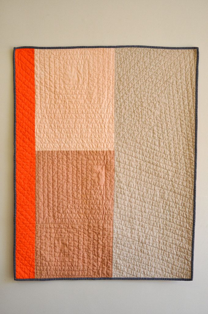 an orange and beige quilt hanging on the wall next to a white wall with a black frame