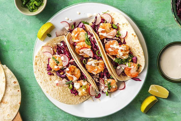 three tacos with shrimp, cabbage and radishes on a white plate next to two glasses of ranch dressing