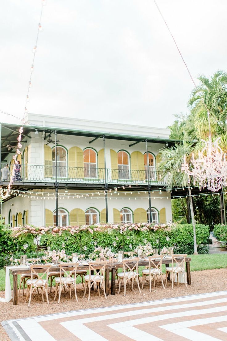 an outdoor dining area with tables and chairs in front of a large white building surrounded by greenery