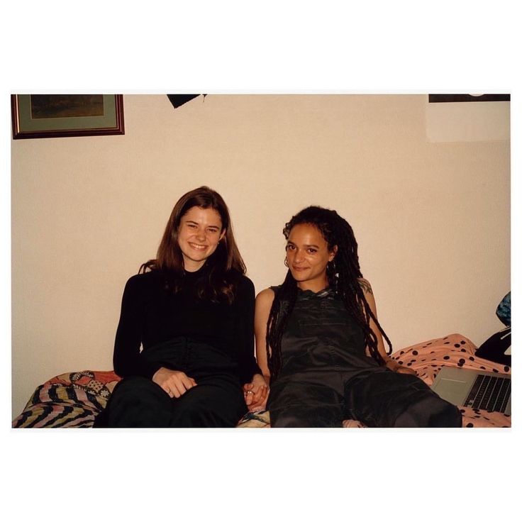 two young women sitting on top of a bed next to each other in front of a white wall