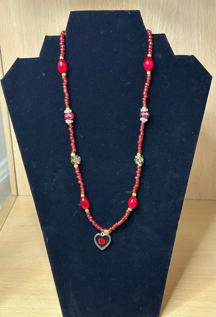 Venture into the unknown with this daring red necklace. Adorned with gold and rhinestones, it showcases a stunning red flower within a heart pendant. Handcrafted with glass beads, a semi-stretch cord, and a metal lobster clasp, this 22-inch piece will captivate and inspire. Spiritual Red Beaded Necklaces With Gold Beads, Spiritual Red Beaded Necklace With Gold Beads, Red Spiritual Jewelry With Gold Beads, Spiritual Red Jewelry With Gold Beads, Adjustable Red Necklace With Heart Beads, Red Adjustable Necklace With Heart Beads, Adjustable Colorful Beads Necklace For Valentine's Day, Red Spiritual Necklace With Heart Beads, Spiritual Red Necklace With Heart Beads