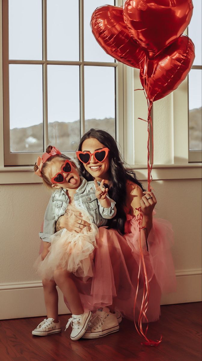 a woman holding a baby wearing sunglasses and sitting in front of a window with red balloons