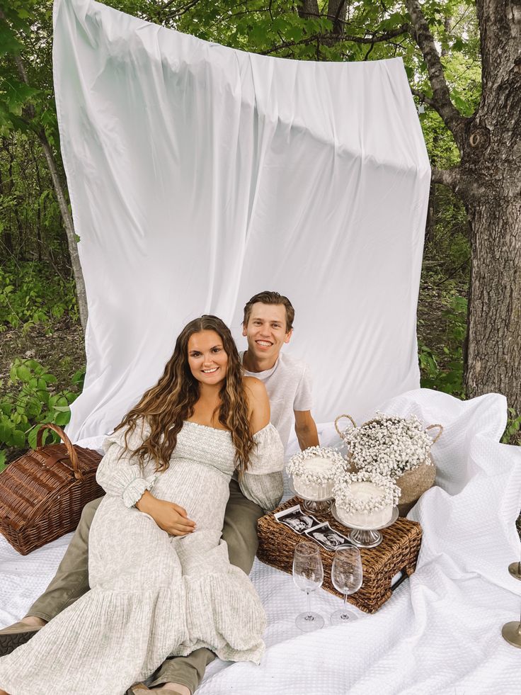 a pregnant couple sitting on a blanket in the woods