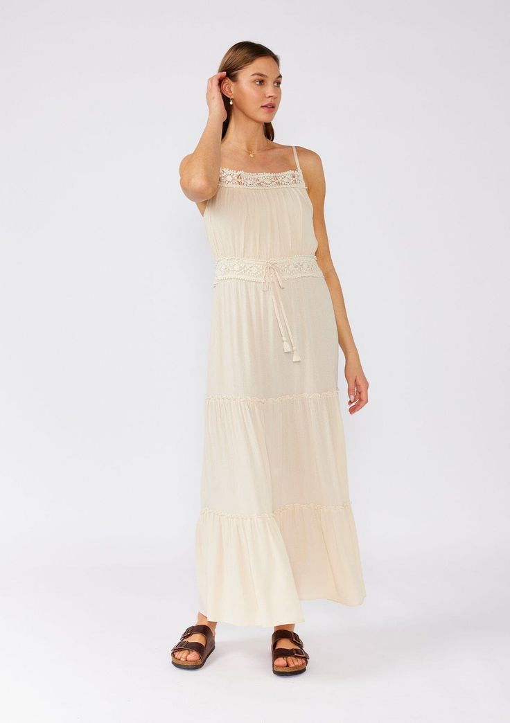 [Color: Natural] A front facing image of a brunette model wearing an off white sleeveless maxi dress. With adjustable spaghetti straps Bohemian Tiered Dress With Adjustable Straps, Bohemian Dress With Adjustable Straps And Tiered Design, Bohemian Spaghetti Strap Dress With Crochet Trim, Sundress Style Maxi Dress With Lace Trim, Spring Tiered Dresses With Crochet Trim, Bohemian Dress With Crochet Trim And Spaghetti Straps, Summer Tiered Maxi Dress With Lace Trim, Vacation Dress With Lace Trim And Tiered Skirt, Summer Maxi Dress With Lace Trim And Tiered Skirt