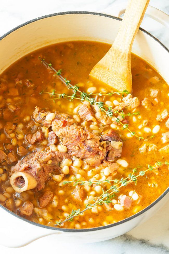 a pot full of soup with meat, beans and herbs on the side next to a wooden spoon