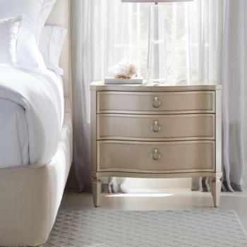 a bedroom scene with focus on the bed and nightstand, which has a white curtained window behind it