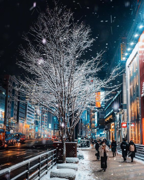 people are walking on the sidewalk in the snow at night time with lights and buildings behind them