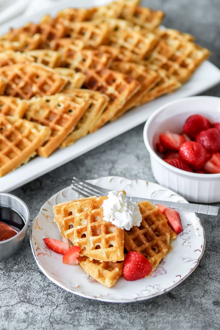 waffles with strawberries and whipped cream are on a plate next to a platter of fruit