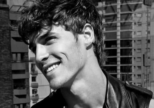 black and white photograph of a young man smiling in front of tall buildings with his hair pulled back