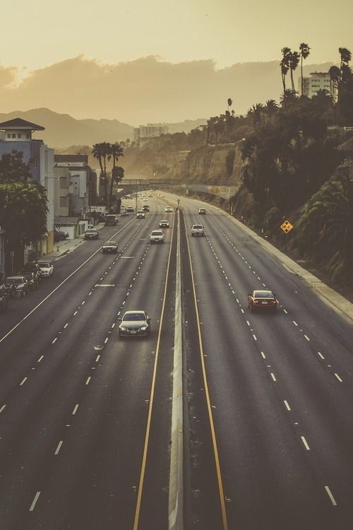 cars are driving down an empty highway in the city at sunset or sunrise, with palm trees and mountains in the background