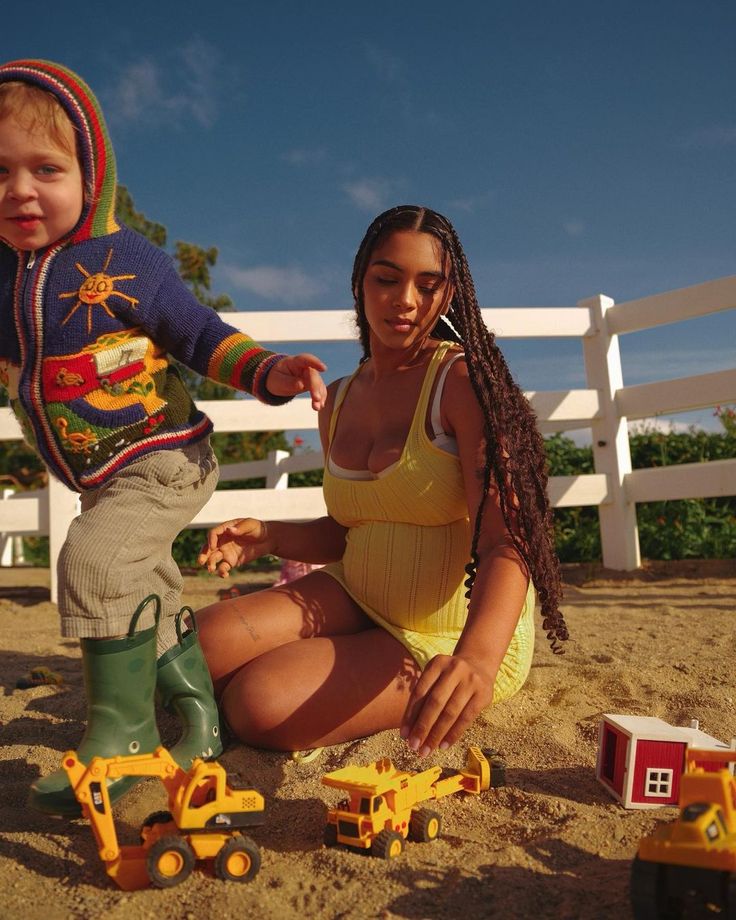 a woman kneeling down next to a child on the beach with toy trucks in front of her