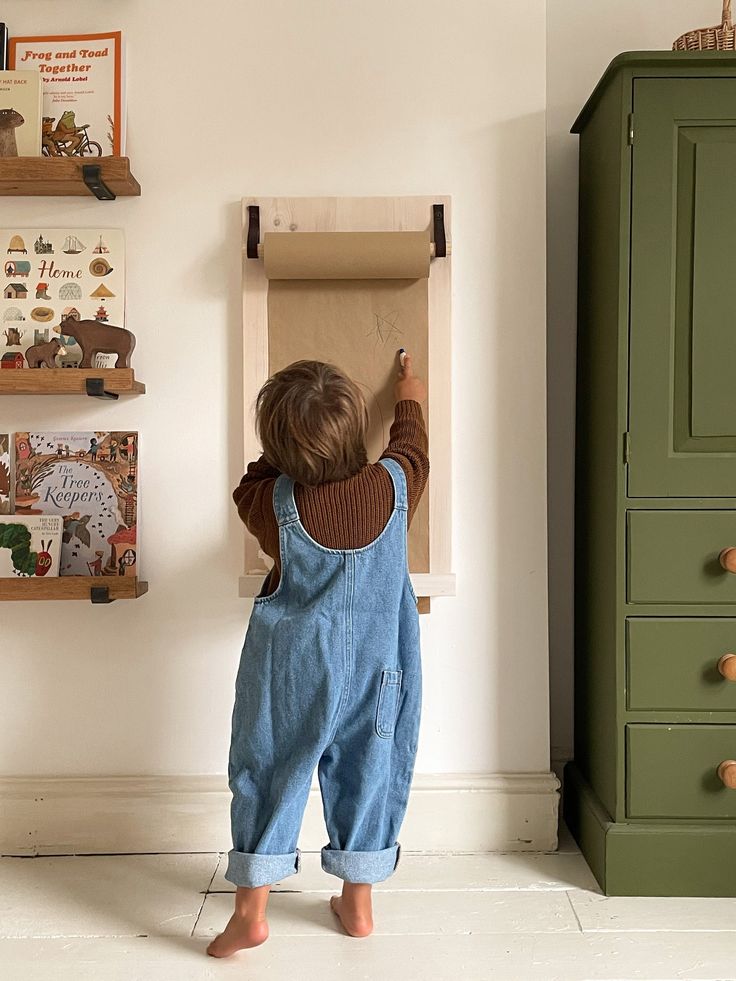 a little boy standing in front of a green cabinet and looking at the wall with pictures on it