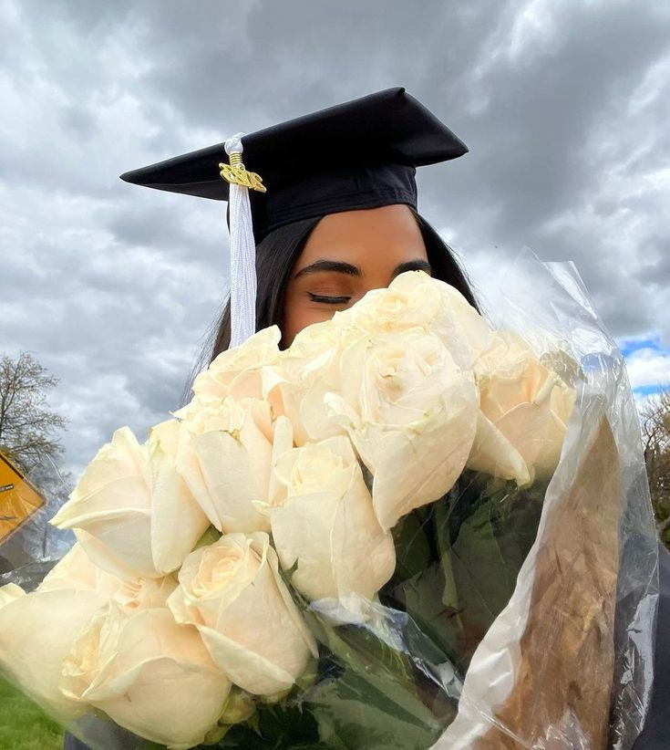 a woman wearing a graduation cap and gown holding flowers in front of her face with a cloudy sky behind her