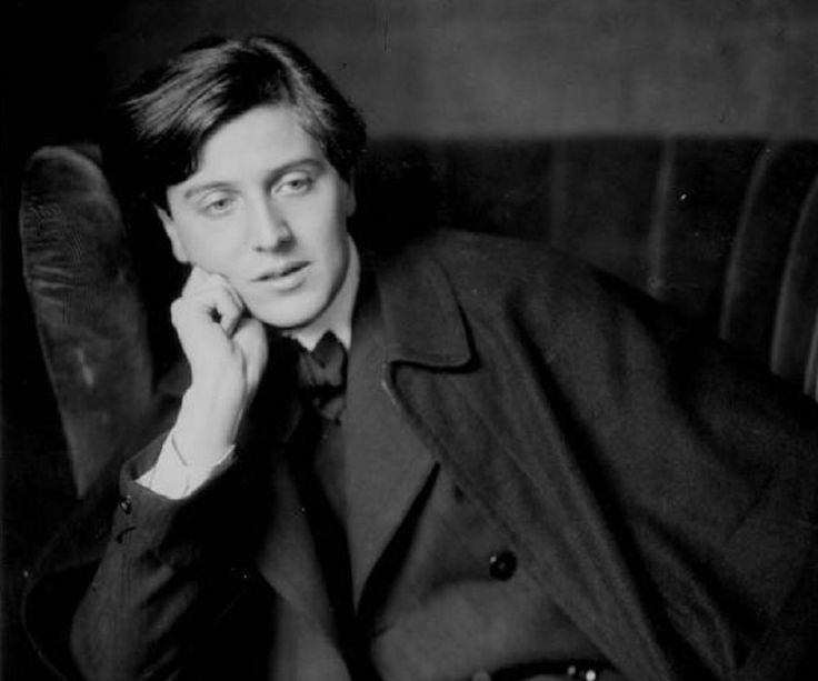 black and white photograph of man in trench coat sitting on chair with hand under chin