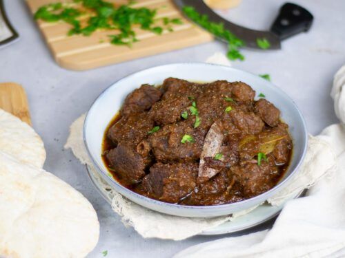 a white bowl filled with meat and vegetables next to some bread on a cutting board