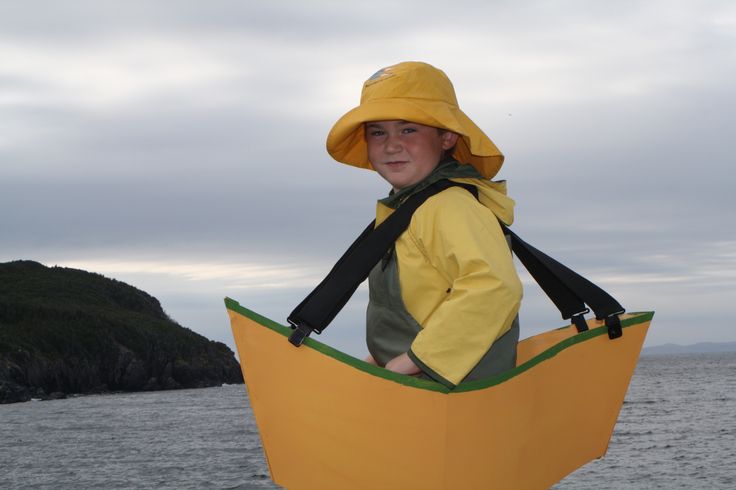 a young boy wearing a yellow hat and carrying a paper boat on the water with an island in the background