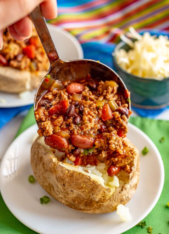 a person scooping chili onto a baked potato on a white plate with green table cloth