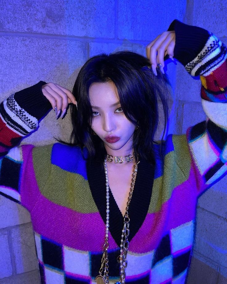 a woman with black hair wearing a multicolored sweater and holding her hands behind her head