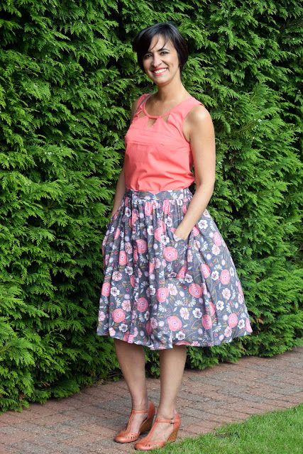 a woman standing in front of a hedge wearing a pink top and floral print skirt